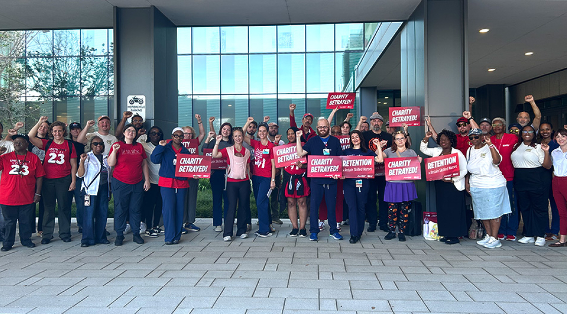 Large group of nurses outside hospital with raised fists, some holding signs "Charity Betrayed"