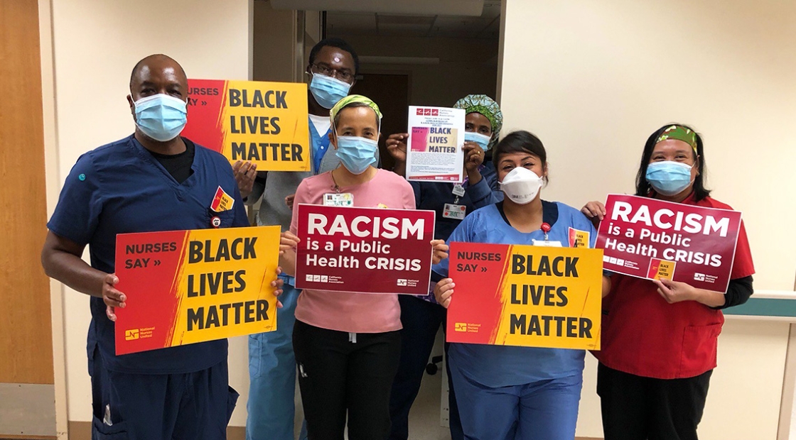 Group of nurses inside hospital hold signs "Nurses Say: Black Lives Matter" and "Racism is a Public Health Crisis"