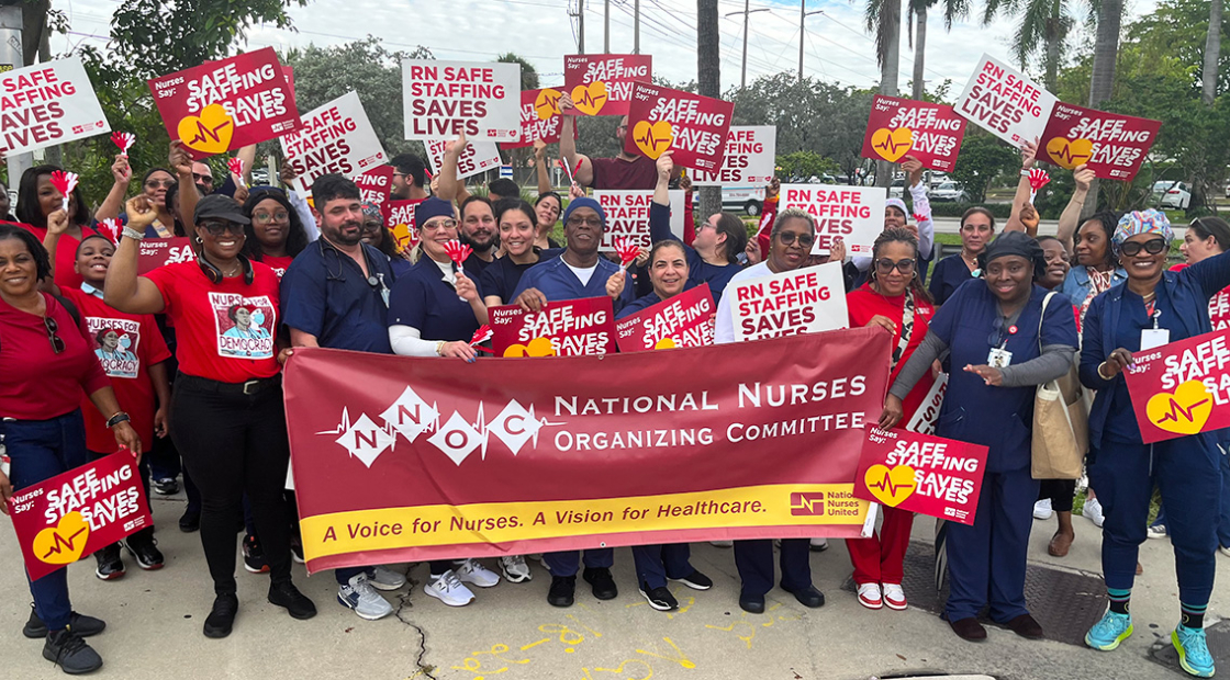 Large group of nurses outside, holding banner "National Nurses Organizing Committee" and signs "Safe Staffing Saves Lives"