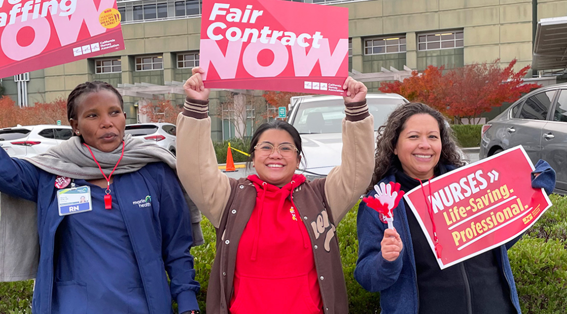 Three nurses outside hospital holding signs "Fair Conract Now" and "Safe Staffing Now"