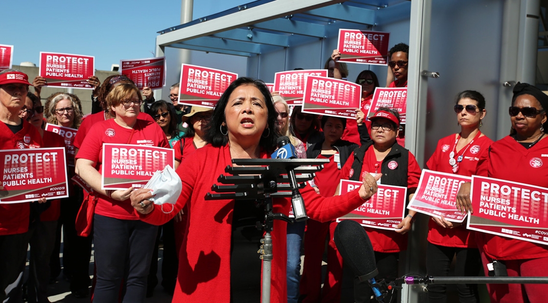 Bonnie Castillo, RN, Executive Director of NNU at podium surrounded by nurses holding signs "Protect Nurses, Patients, Public Health"