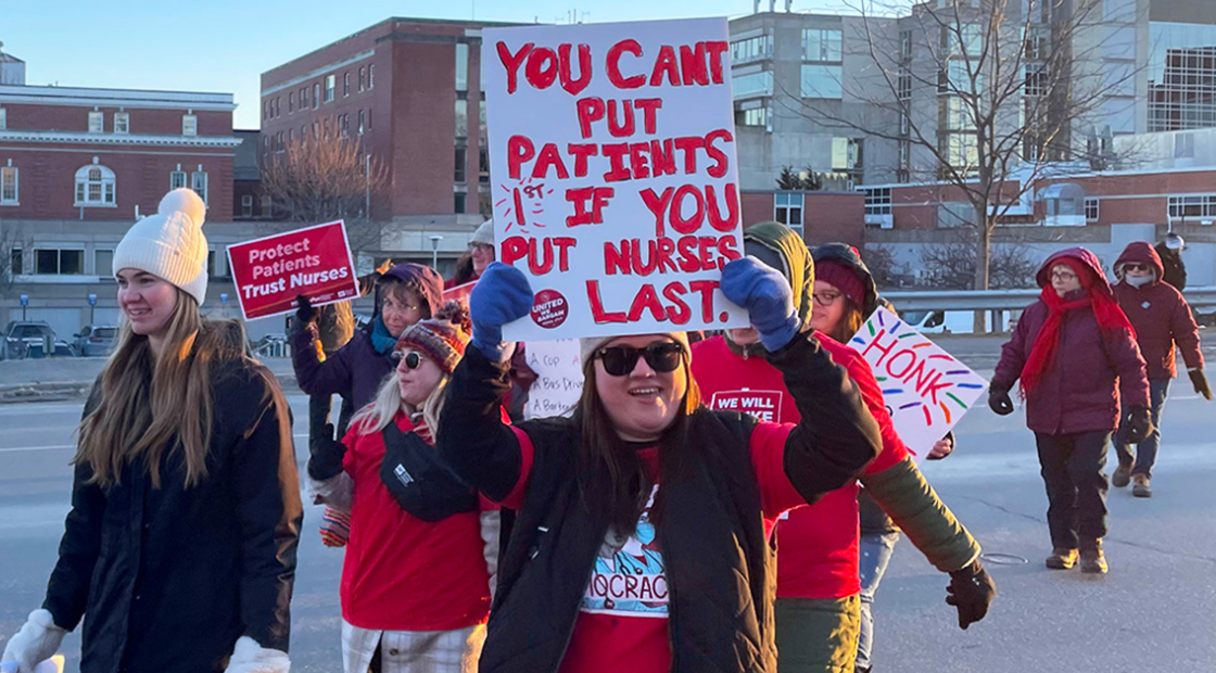 Nurses walking outside of hospital, one holding sign "You can't put patients first if you put nurses last"