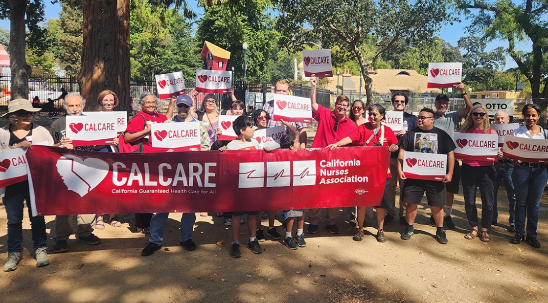 Large group of activists in park holding CalCare signs and banner