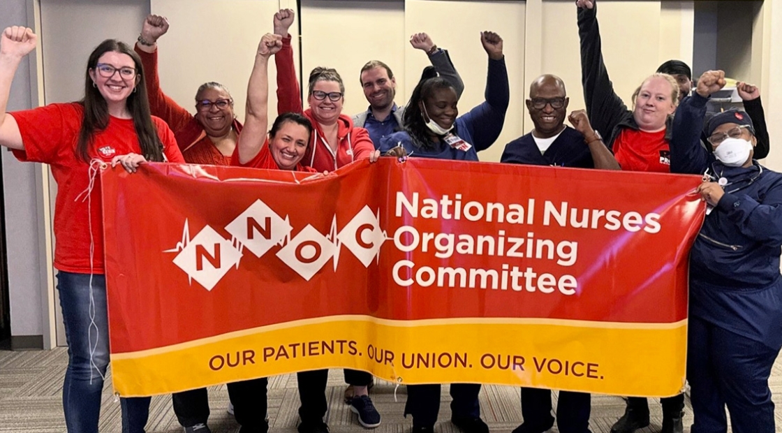Group of nurses smiling, with raised fists, holding banner "National Nurses Organizing Committee - Our Patiens, Our Union, Our Voice""