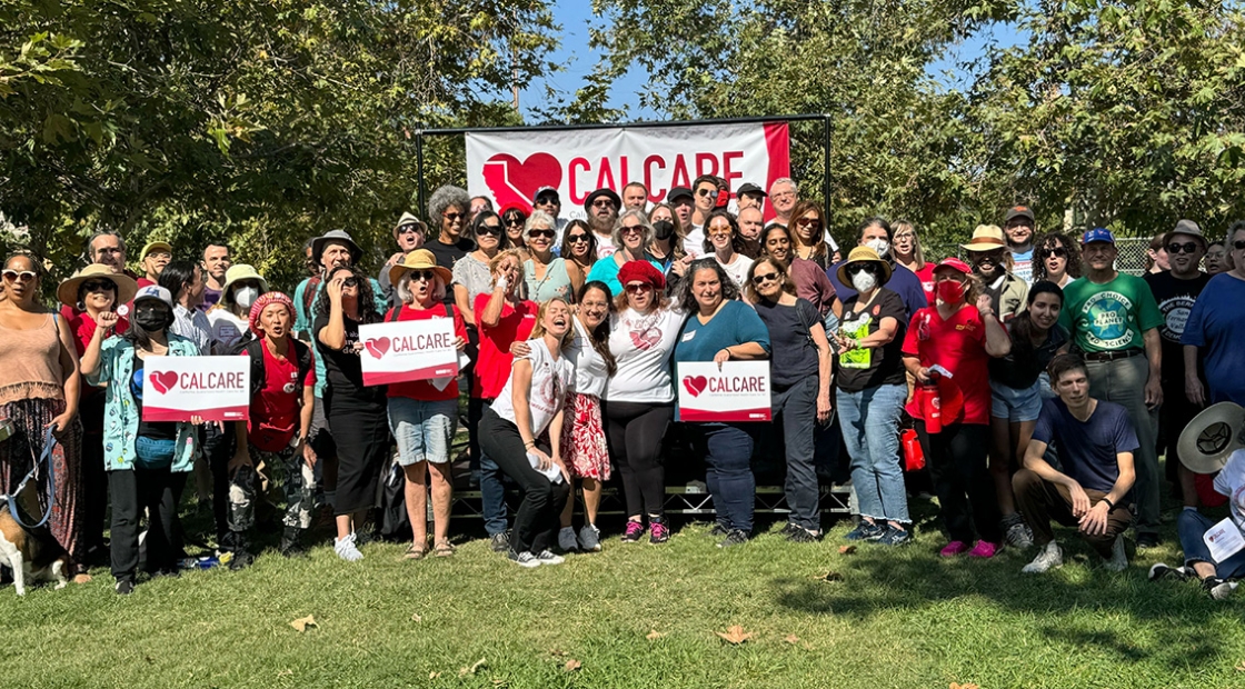 Large group of people in park standing and sitting in front of CalCare banner, some holding CalCare signs