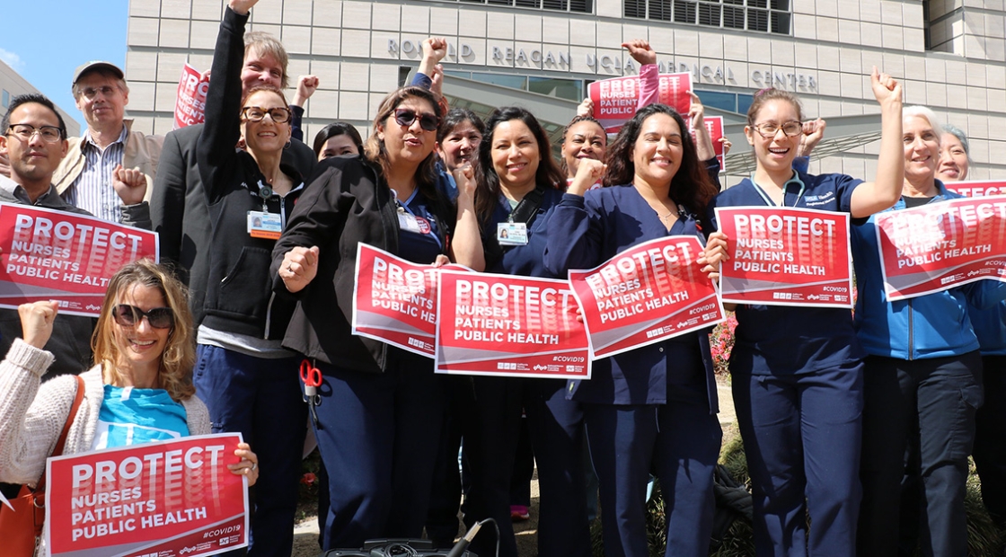 Group of nurses with raised fists, holding signs "Protect Nurses, Patients, Public Health"