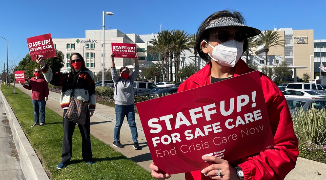 Four nurses standing outside hospital holding signs "Staff Up for Safe Patient Care"