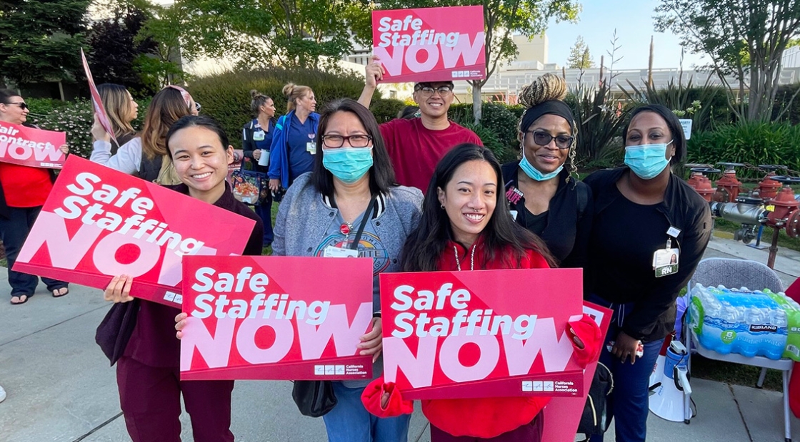 Group of nurses outside, smiling, holding signs "Safe Staffing Now"