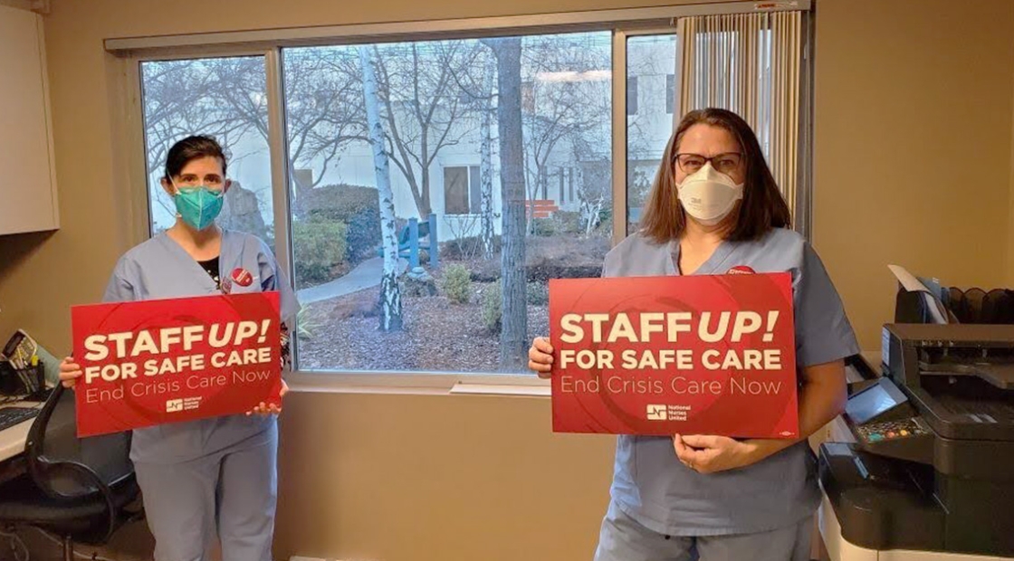 Two nurses inside hospital hold signs "Staff Up For Safe Care"