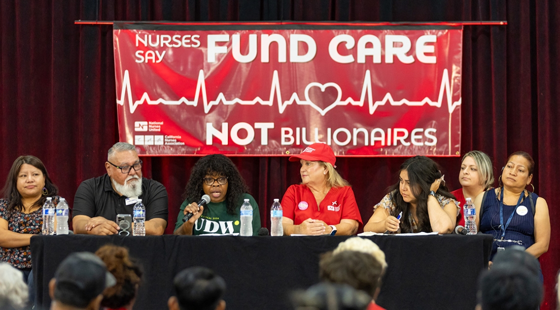 Panel at town hall in front of banner "Fund Care Not Billionaires"