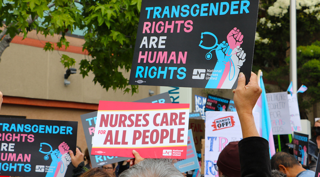 Hands holding signs "Transgender Rights Are Human Rights" and "Nurses Care for All People"