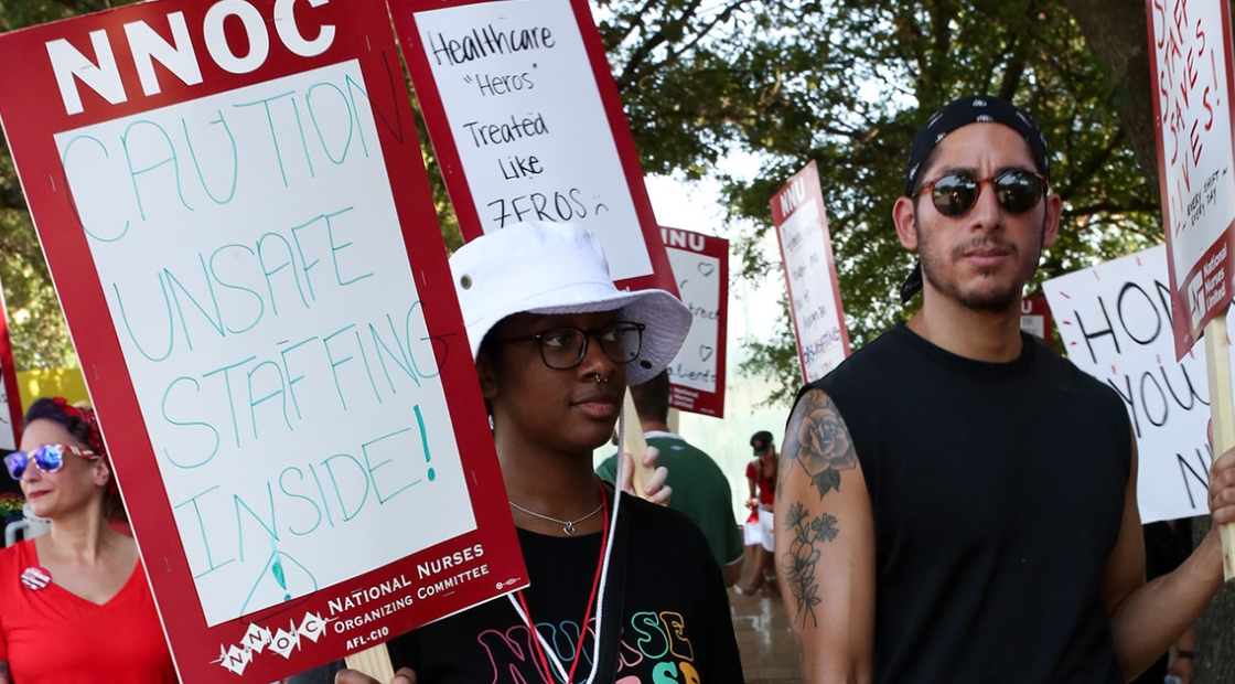 Nurses on picket line, one holds sign "Caution, Unsafe Staffing Inside"