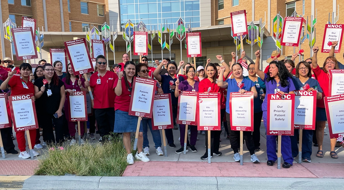 Large group of nurses outside hospital with fists raised, signs that read "Patients are our top priority"
