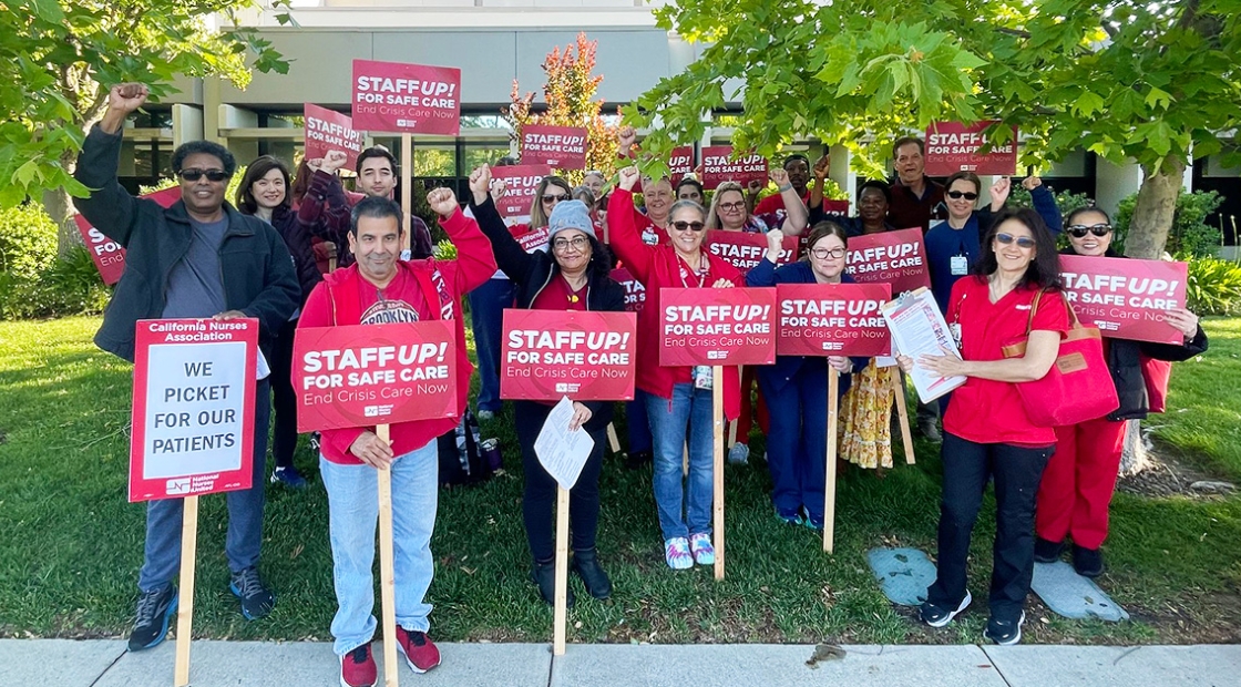 Large group of nurses standing outside hospital with raised fists, holding signs "Staff Up for Safe Patient Care"