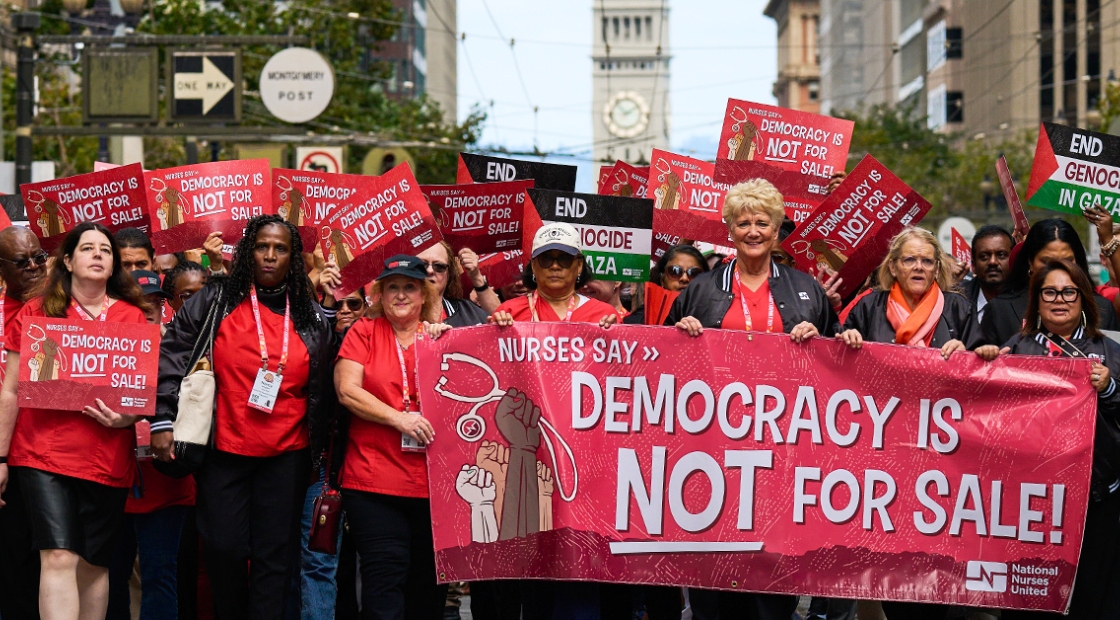 Large group of nurses marching, holding banner "Democracy is NOT for Sale"