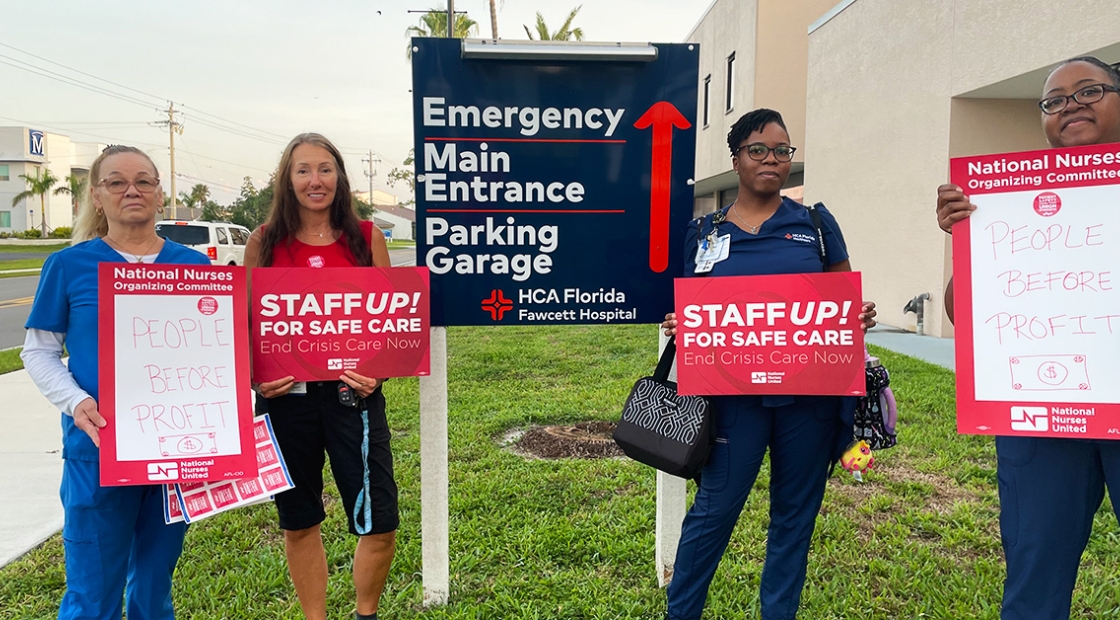 Group of four nurses outside hospital hold signs "People Before Profit" and "Staff Up for Safe Patient Care"