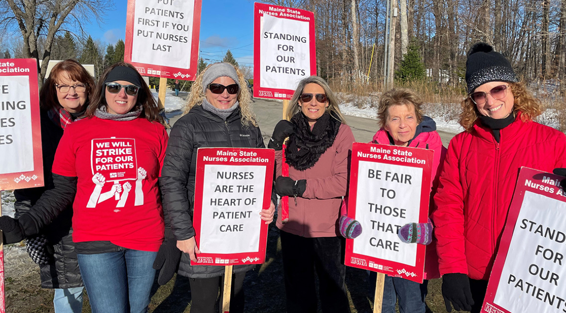 Group of nurses outside dressed in winter clothes, holding signs "Standing for our patients", "Nurses are the heart of patient care", more