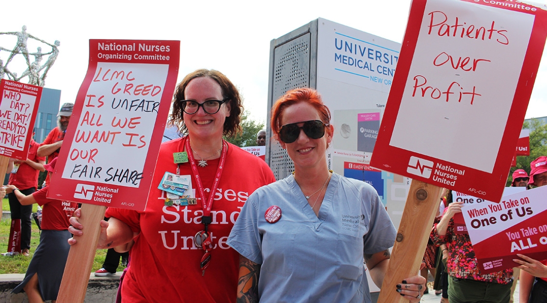 Two nurses smiling, holding signs "LCMC Greed Is Unfair, All We Want Is Our Fair Share" and "Patients Over Profit"