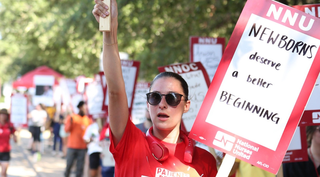 Nurse holding sign "NNU: Newborns deserve a better beginning" and shirt "Patients before profits"