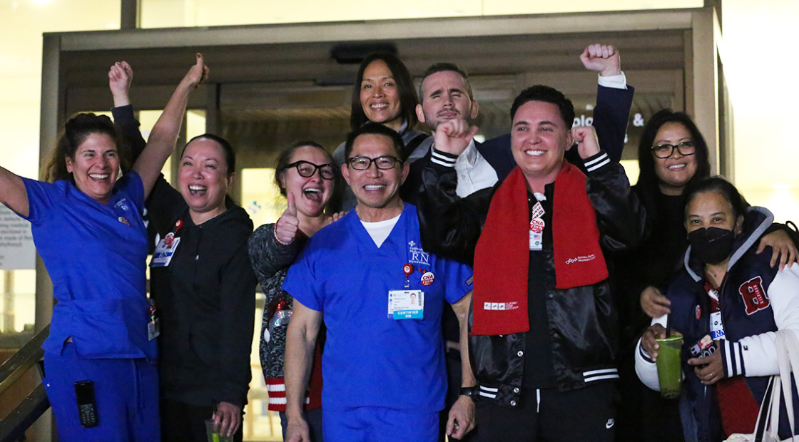 Large group of nurses outside hospital, smiling, with raised fists