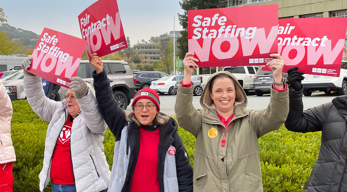 Nurses outside hospital holding signs "Safe Staffing Now" and "Fair Contract Now"