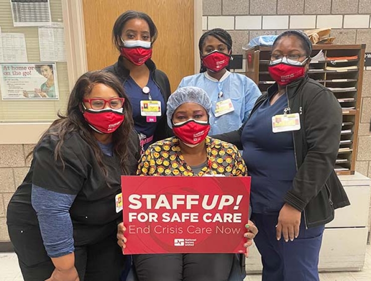 Nurses inside hospital, one holds sign "Staff Up: End Crisis Care"