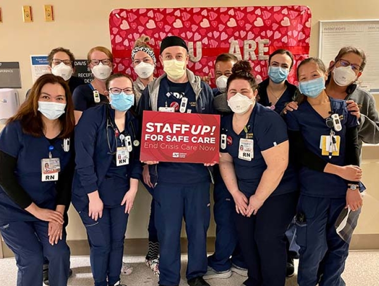 Nurses inside hospital, one holds sign "Staff Up: End Crisis Care"