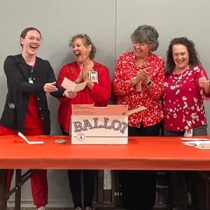 Four nurses standing at table with ballot box, smiling and laughing