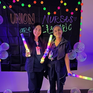 Two nurses smiling and holding glod sticks in front of banner "Union Nurses are Electric"