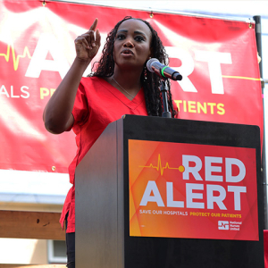 Nurse speaking at podium with banner behind "Red Alert: Save Our Hospitals Tour"