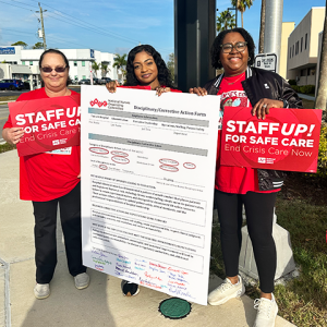 Three nurses holding signs "Staff up for safe care: End crisis care now" and a huge disciplinary/corrective action form