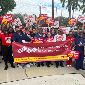 Large group of nurses outside, holding signs calling for safe staffing and NNOC banner
