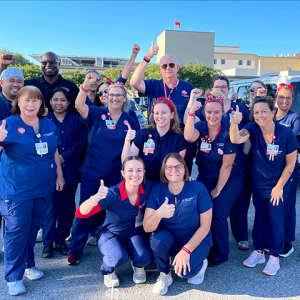 Large group of nurses outside hospital, wearing blud scrubs, smiling, and giving thumbs up