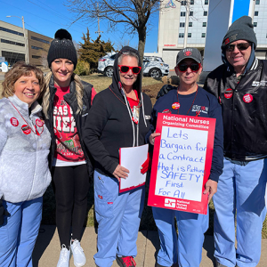 Five nurses outside hospital. One holds sign "Let's bargain for a contract that is putting SAFETY FIRST for all"
