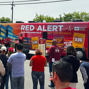 People speak at podium in front of Red Alert Tour bus, crowd watching