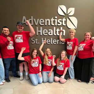 Group of nurses inside, in front of sign "Adventist Health", smiling, wearing red, and with raised fists