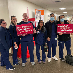 Group of sixe nurses inside hospital, side-by-side, smiling, holding signs "Union Nurse Power"