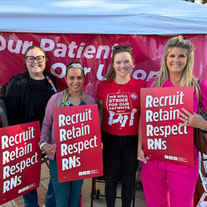 Four nurses, outside, smiling, side-by-side, holding signs "Recruit, Retain, Respect RNs"