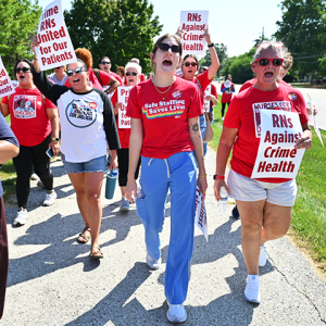 Community and nurses marching and holding signs "RNs Against Crime Health." Person with shirt "Safe Staffing Saves Lives"