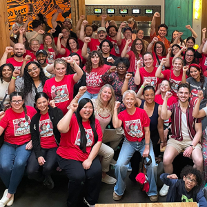 Large group of nurses inside, mostly wearing red, smiling, with raised fists