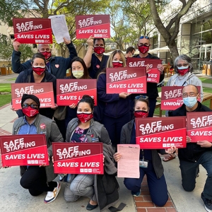 Large group of nurses outside hospital hold signs "Staff Up for Safe Care"