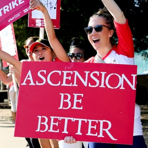 Nurse on picket line holds sign "Ascension Be Better"