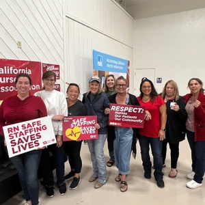 Group of 9 nurses smiling, one holds signs "Respect Nurses, Our Patients, Our Communitites