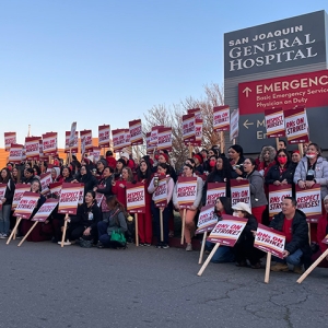 Large group of nurses outside San Joquin General Hospital, smiling and holding signs "RNs On Strike" and "Respect Nurses"