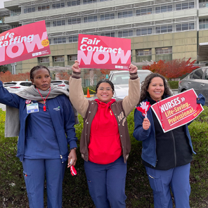 Three nurses outside hospital holding signs "Fair Conract Now" and "Safe Staffing Now"
