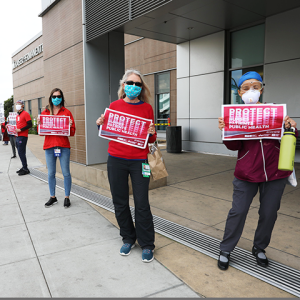 Nurses outside hospital hold signs "Protect Nurses, Patients, Public Health"