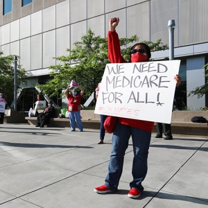Nurse raising fist and holding sign "We need Medicare For All!"