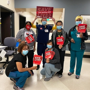 Group of four nurses inside hospital hold signs "Safe Staffing Now", "Save Lives"