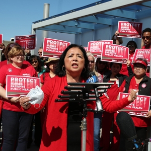 Bonnie Castillo, RN, Executive Director of NNU at podium surrounded by nurses holding signs "Protect Nurses, Patients, Public Health"