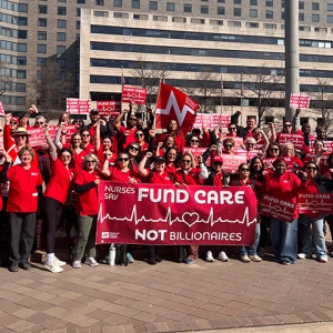 Large group of nurses outside holding banner and signs "Nurses Say: Fund Care Not Billionaires"